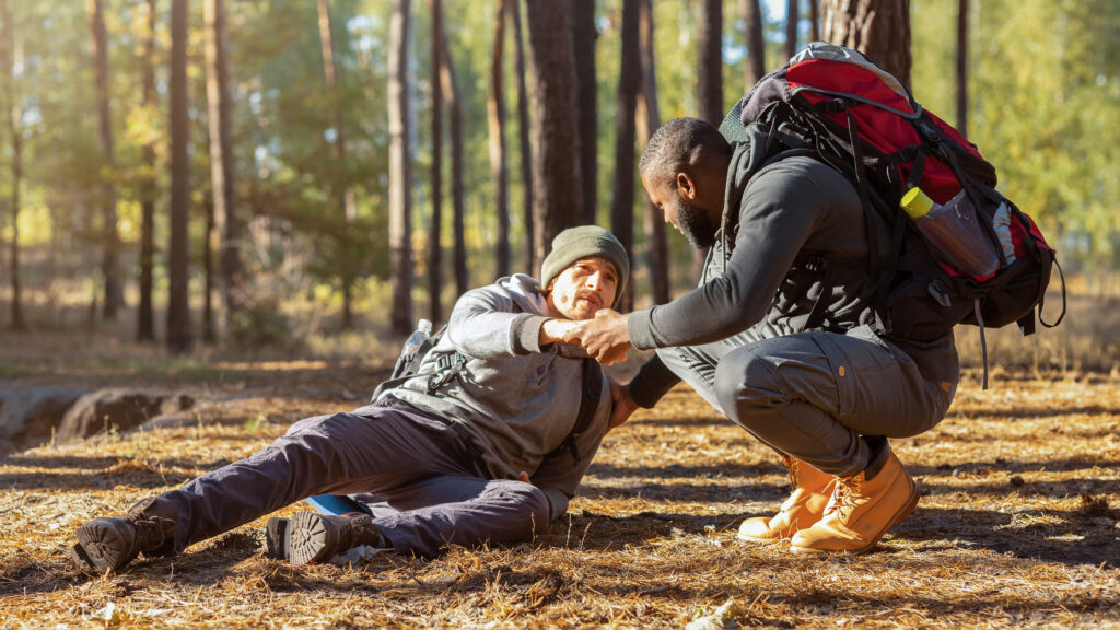 African guy helping his injured friend to get up while hiking, copy space